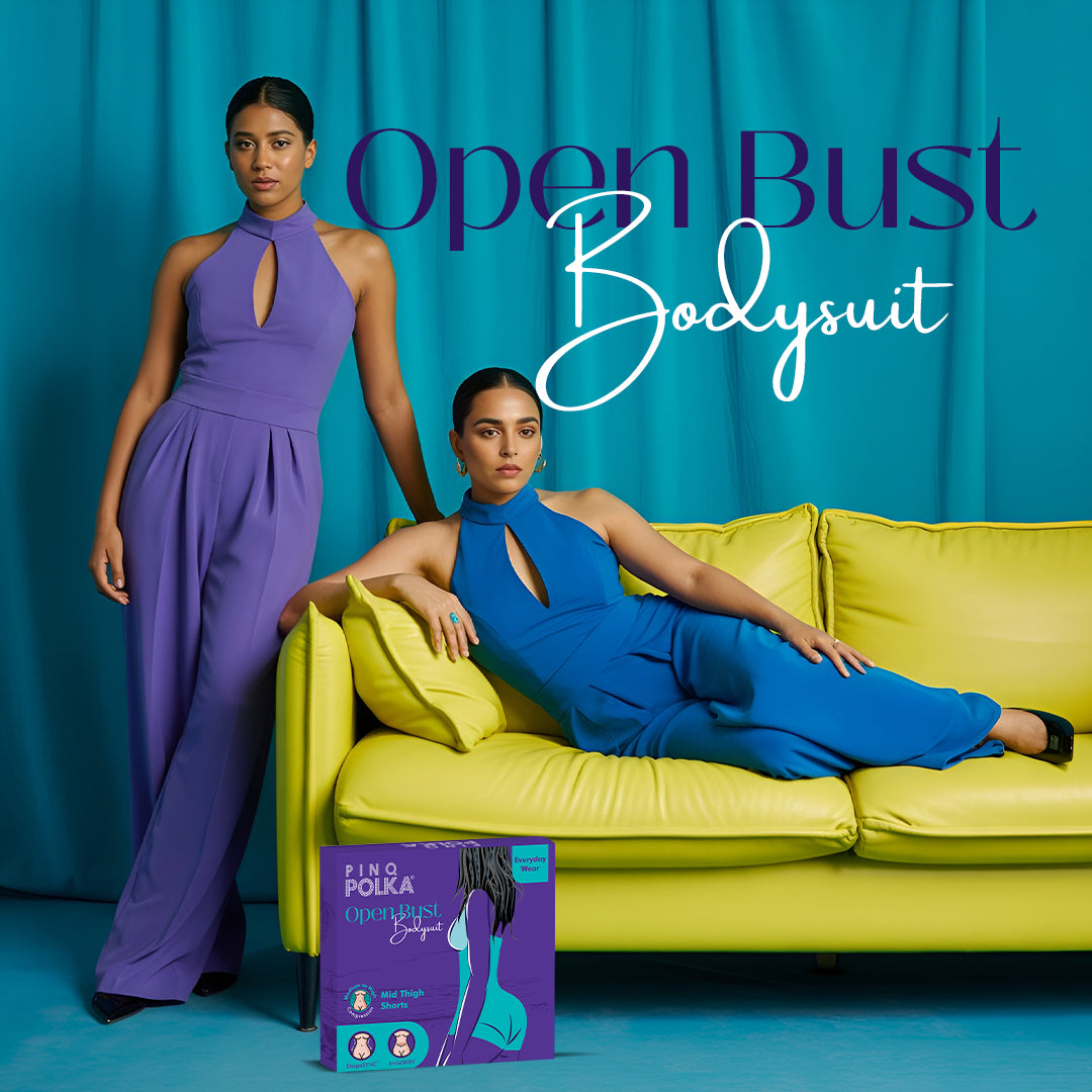 Two women wearing open bust bodysuits with a blue curtain and yellow couch in the background.